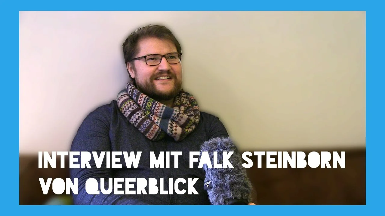 Ein lächelnder Mann mit Brille und buntem Schal hält ein Plüschtier, dazu Text "Interview mit Falk Steinborn von Queerblick".