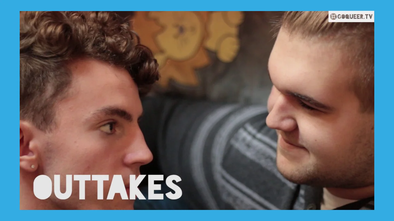 Zwei Männer blicken sich lächelnd an, im Hintergrund ein unscharfes Bild, Text "OUTTAKES" eingeblendet.