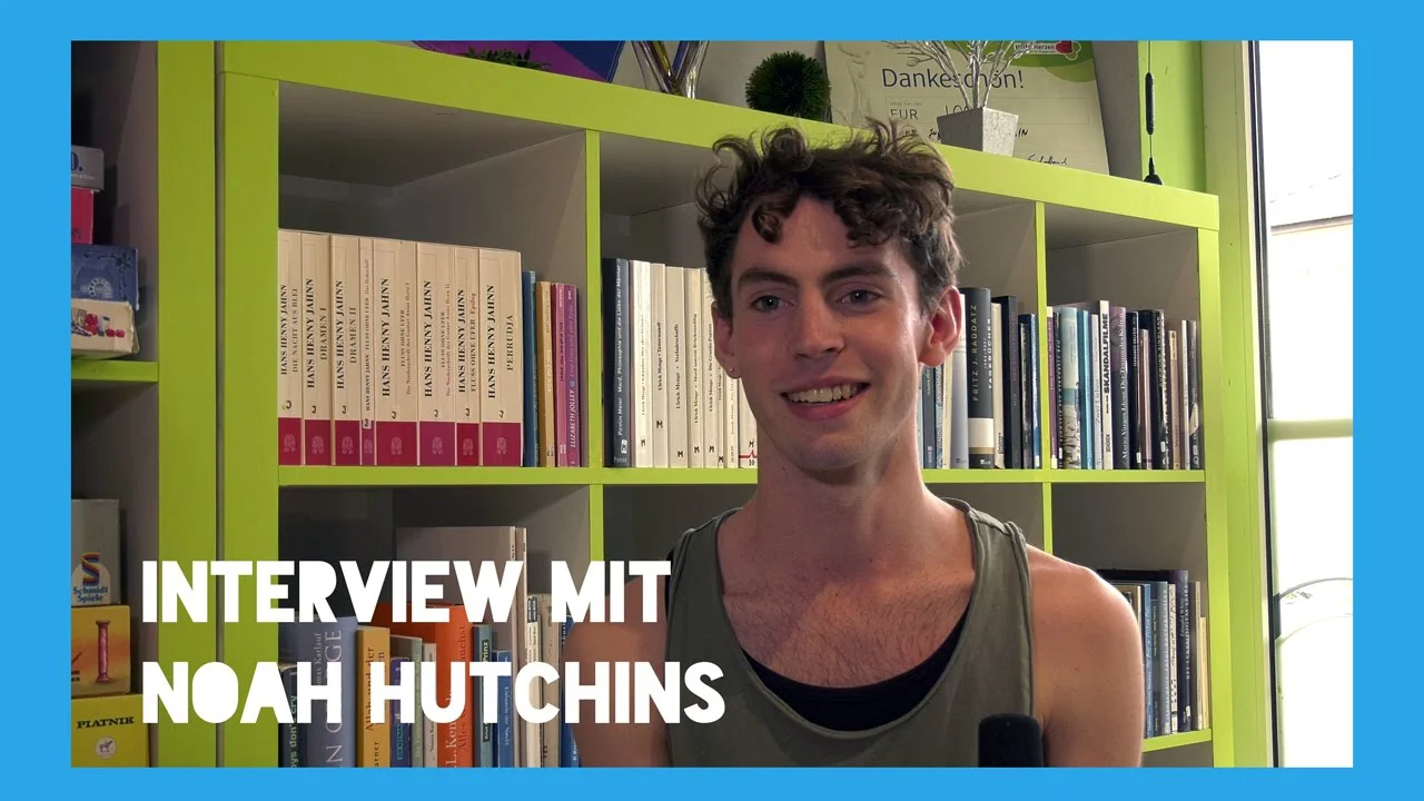 Junger Mann mit lockigem Haar steht vor einem grünen Bücherregal, Text "Interview mit Noah Hutchins" eingeblendet.