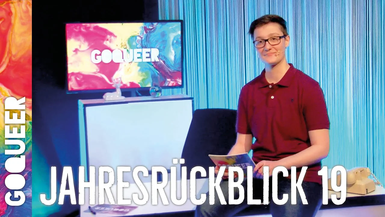Junger Mann mit Brille und Poloshirt steht vor einem Schreibtisch mit Monitor, auf dem "GOQUEER" zu sehen ist.
