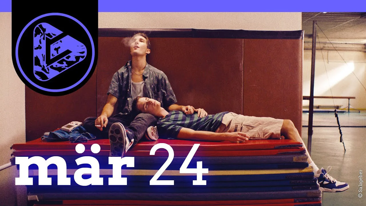 Zwei Personen auf gestapelten roten Matratzen, eine sitzend, eine liegend, vor beiger und roter Wand.