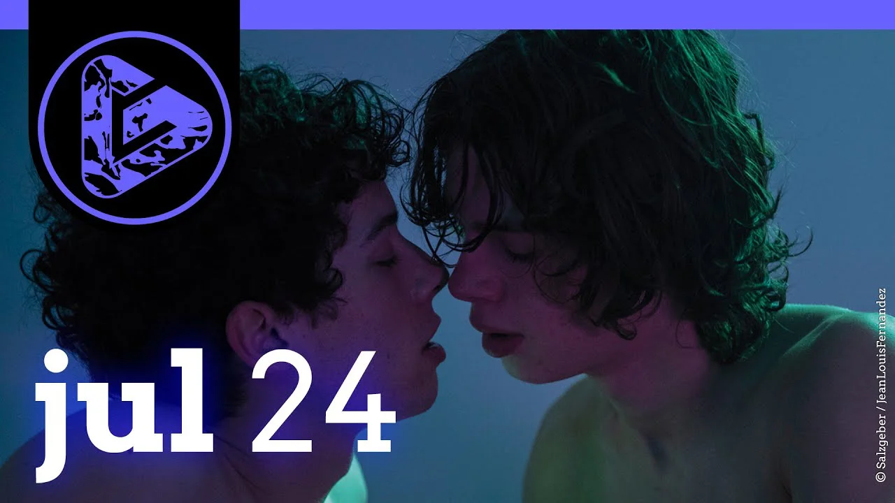 Zwei junge Menschen mit dunklen lockigen Haaren stehen sich sehr nahe gegenüber vor blauem Hintergrund.