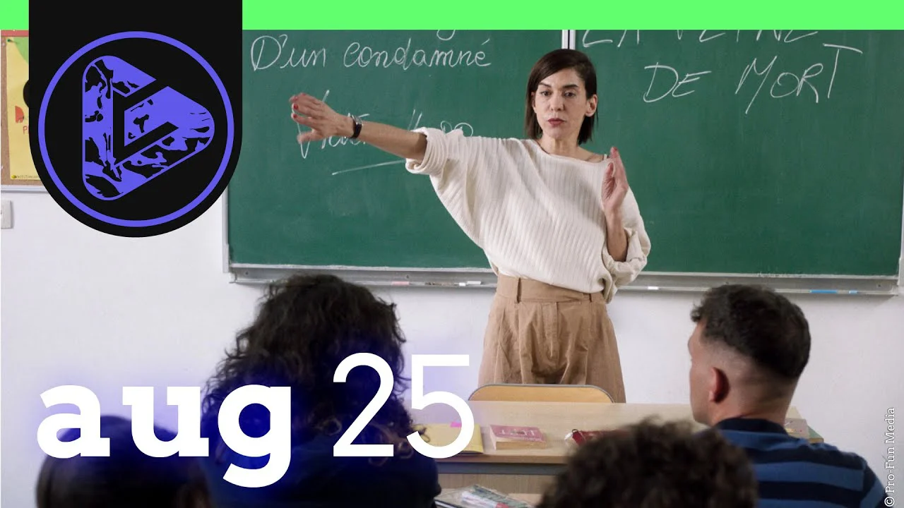 Lehrerin steht vor Tafel in Klassenzimmer und erklärt etwas, Schüler sitzen davor, Logo links oben.